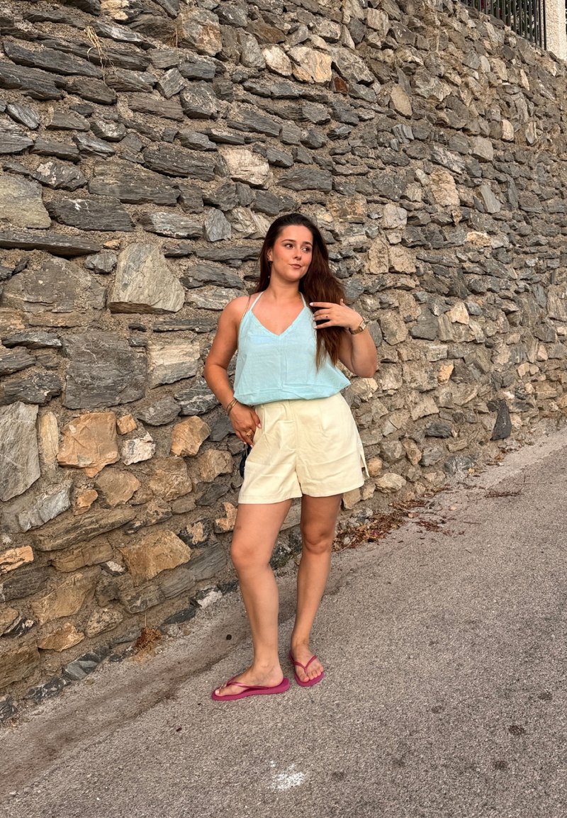 Jeune femme portant un haut bleu clair, un short crème et des tongs roses, debout sur un chemin pavé à côté d'un mur en pierre rugueuse.