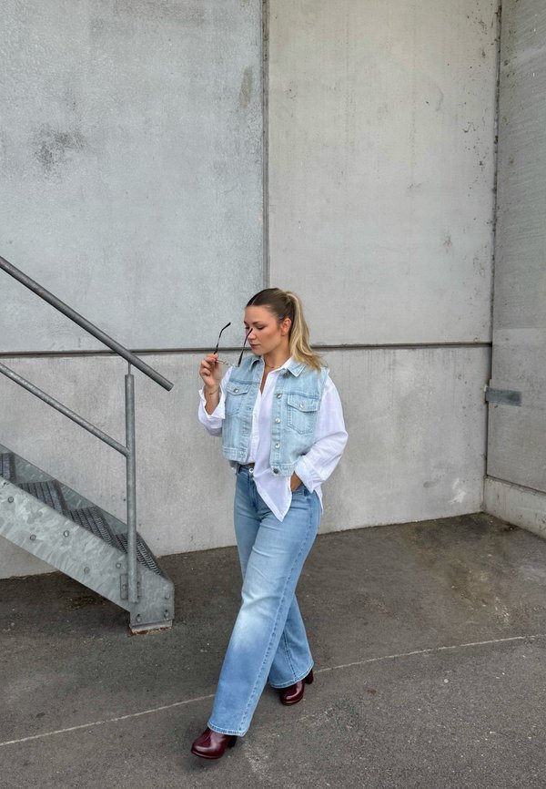 Woman in light denim vest and jeans holding sunglasses walking near metal stairs against a concrete wall background.
