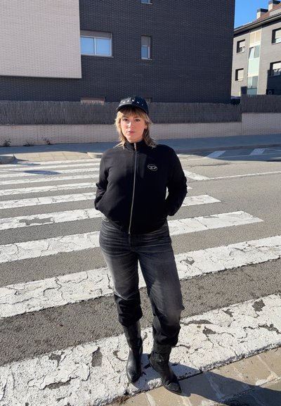 Chaqueta negra con cremallera y logo, jeans grises y botas negras hasta la rodilla. De pie en un paso de peatones con un fondo de asfalto texturizado.