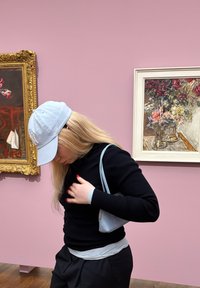 Gai zils cepure, melns ribots garām piedurknēm tops, pelēks slāņots krekls un melnas bikses. Maza pelēka plecu soma, stāvot blakus gleznai.