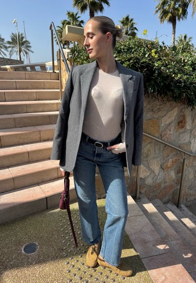 Mujer con blazer gris, blusa beige claro, jeans azules y zapatos color marrón claro parada cerca de unas escaleras al aire libre, sosteniendo un bolso pequeño y oscuro.