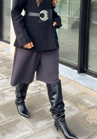 Giacca nera su misura, pantaloni shorts grigi a gamba larga, e stivali alti neri in pelle con punta sottile e texture arricciata. Fibbia circolare in argento.
