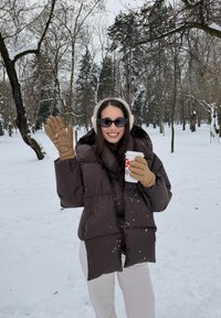 Femme portant des lunettes de soleil, des cache-oreilles, une doudoune brune et des gants, tenant un gobelet de café à emporter, faisant un signe de la main avec une forêt enneigée en arrière-plan.