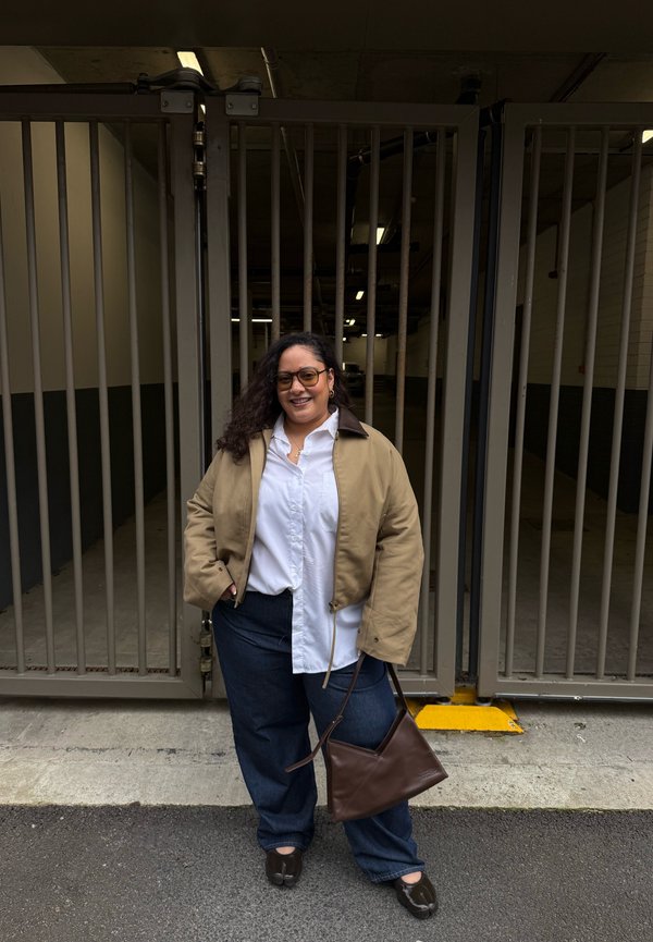 Mujer con cabello rizado, gafas, chaqueta beige, camisa blanca y pantalones azules, sonriendo con las manos en los bolsillos junto a una puerta metálica.