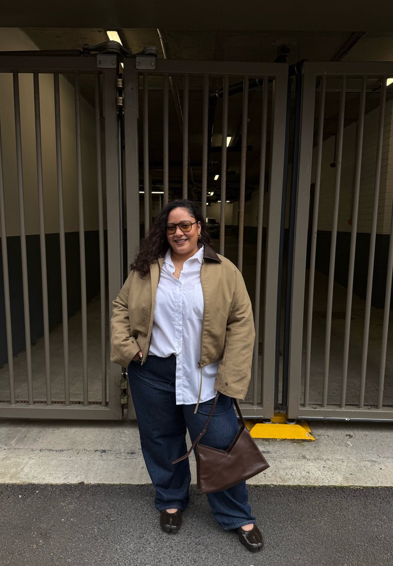 Femme aux cheveux bouclés portant des lunettes, une veste beige, une chemise blanche et un pantalon bleu, souriant avec les mains dans les poches près d'une grille métallique.