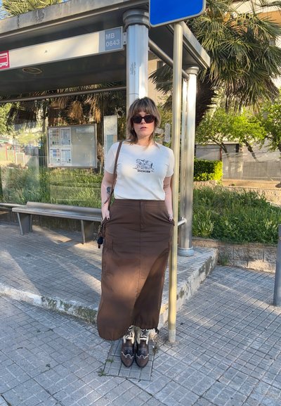 Larga falda marrón con bolsillos, camiseta beige con estampado gráfico y gafas de sol oscuras. Superficies texturizadas y fondo urbano.