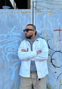 White canvas jacket over a gray hoodie, standing against a rusted metal backdrop with graffiti. Black sunglasses, light beige pants, and keys visible.