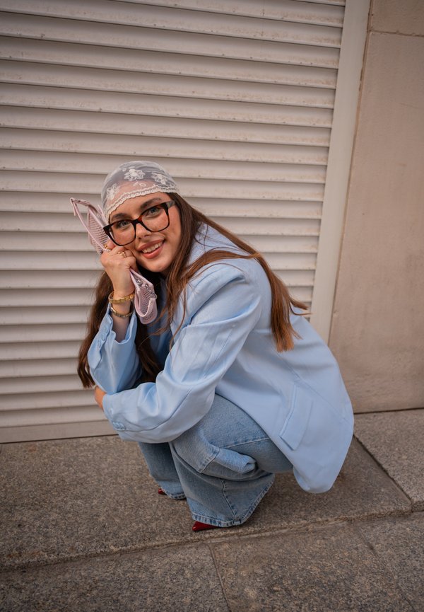 Una mujer con gafas y un pañuelo de encaje en la cabeza se agacha junto a una persiana, sonriendo mientras sostiene un bolso rosa contra su mejilla.