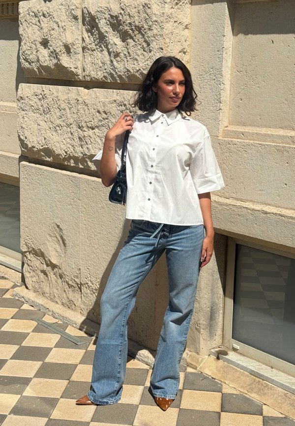 Mujer con camisa blanca abotonada, vaqueros azules y zapatos marrones puntiagudos, de pie contra una pared de piedra texturizada a la luz del sol.