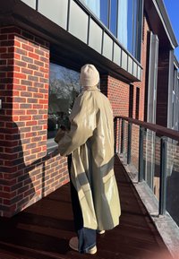 Une personne portant un manteau beige brillant et un bonnet en laine se tient sur un balcon, faisant face à un bâtiment en briques aux grandes fenêtres, par une journée ensoleillée.