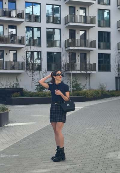 Mujer con gafas de sol y ropa negra parada en una zona pavimentada cerca de un edificio moderno de apartamentos con balcones y árboles pequeños.