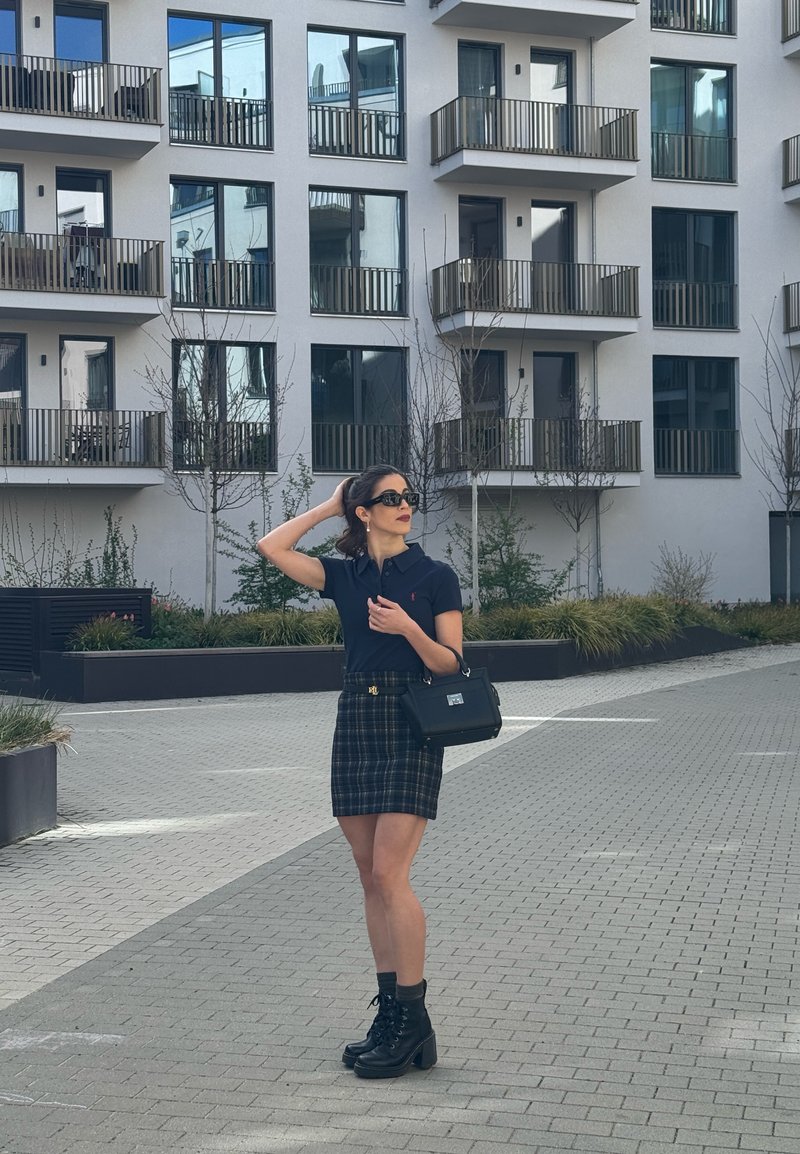 Donna con occhiali da sole e abbigliamento nero in piedi su un'area pavimentata vicino a un moderno edificio di appartamenti con balconi e piccoli alberi.