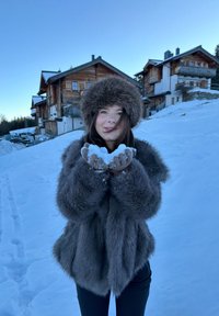 Jeune femme portant un manteau de fourrure sombre et un chapeau, tenant de la neige dans des mains gantées, avec des chalets en bois et une colline enneigée en arrière-plan.