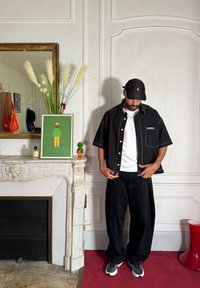 Μαύρο κοντομάνικο πουκάμισο με κουμπιά και αντίθετη ραφή, λευκό μπλουζάκι από κάτω, φαρδιά μαύρα παντελόνια και μαύρα αθλητικά παπούτσια.