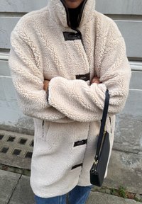 Manteau en polaire de couleur crème avec un col montant, doté de fermetures à bascule foncées et de poches latérales. Porté avec un jean bleu et un sac noir.