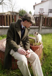 Mladý muž v olivovo zelenom kabáte a plochej čiapke sedí pri kvetináči. Biela mačka s čiernymi akcentmi sa približuje, obklopená trávou.