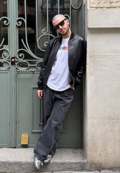 Chaqueta bomber de cuero negra sobre una camiseta blanca, jeans de pierna ancha grises y zapatos plateados brillantes, posando frente a puertas verdes ornamentadas.