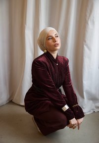 Outfit borgogna con texture, maniche lunghe e design aderente, abbinato a un hijab beige e accenti di gioielli dorati. Sfondo neutro.
