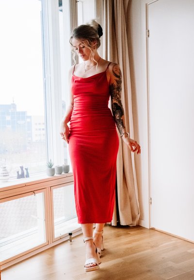 Vestido rojo ajustado con tirantes finos y textura fruncida, que llega a la mitad de la pantorrilla. Combinado con zapatos de tacón ligeros y con tiras. Fondo de suelo de madera natural.