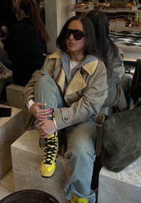 Trench-coat beige avec un sweat à capuche gris, jean bleu clair et baskets jaune vif. Assis sur une surface en pierre claire dans un cadre de café.