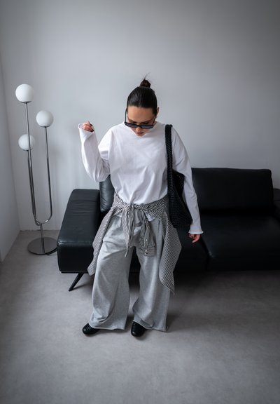Persona que lleva una camisa blanca de manga larga de gran tamaño, pantalones grises de pierna ancha, camisa de rayas atada a la cintura, zapatos negros, gafas de sol y sostiene un bolso negro en interiores.