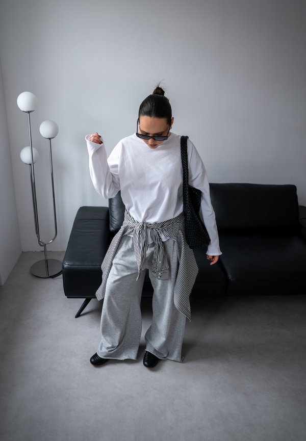 Personne portant une chemise blanche à manches longues surdimensionnée, un pantalon gris à jambes larges, une chemise rayée nouée à la taille, des chaussures noires, des lunettes de soleil et tenant un sac noir à l'intérieur.