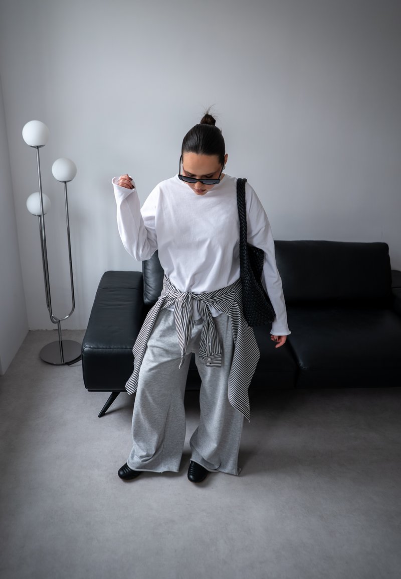 Persona valkā pārāk lielu baltu ar garām piedurknēm kreklu, pelēkas plaša piegriezuma bikses, svītrains krekls sasiets jostasvietā, melnas kurpes, saulesbrilles un tur melnu somu telpās.