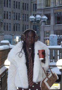 Jeune femme en manteau de fourrure blanc et cache-oreilles léopard tenant une tasse de café et un sac en peau retournée marron dans un cadre urbain enneigé.
