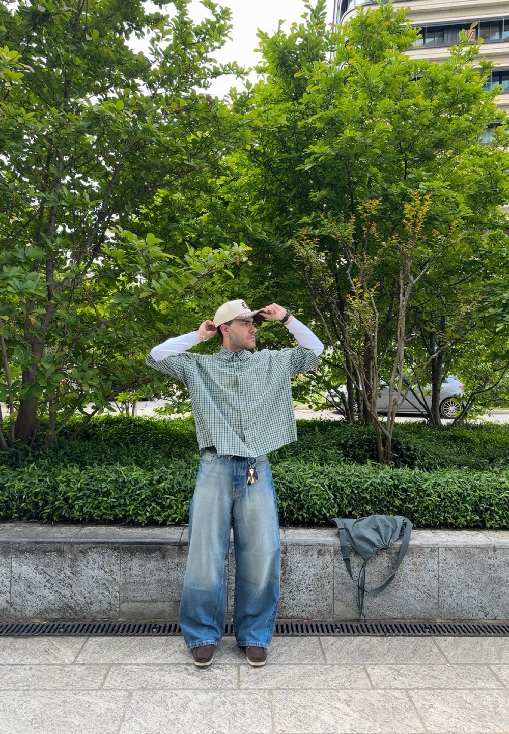 Person i en grön och vit rutig oversized kortärmad skjorta, lösa blå jeans med en urtvättad textur och bruna skor, står vid grönska.