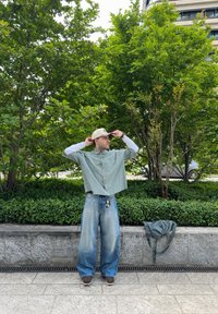 Person i en grön och vit rutig oversized kortärmad skjorta, lösa blå jeans med en urtvättad textur och bruna skor, står vid grönska.