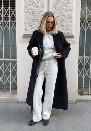 Mujer con cabello rubio, gafas, sudadera con capucha gris y pantalones deportivos, abrigo largo negro, sosteniendo una taza de café y un bolso negro, de pie al aire libre.