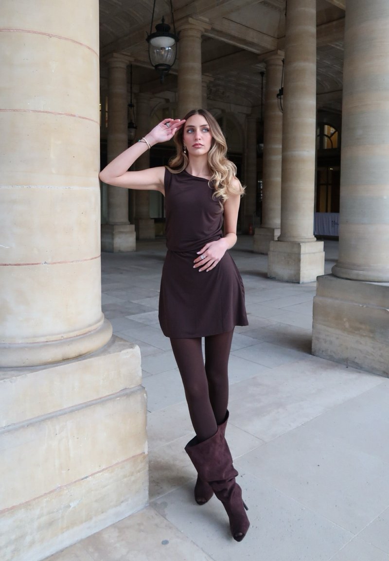 Γυναίκα με μακριά κυματιστά μαλλιά ποζάρει δίπλα σε πέτρινες κολόνες, φορώντας αμάνικο καφέ φόρεμα, ταιριαστά κολάν και χαλαρά σκούρα μπότες μέσα σε εσωτερικό χώρο.