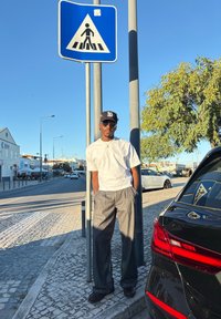 Un bărbat îmbrăcat într-un tricou alb și pantaloni gri cu dungi stă lângă un semn de trecere pentru pietoni, cu o mașină neagră vizibilă în spatele său.