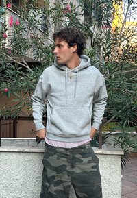 Hanorac gri din fleece cu buzunar tip kangaroo și glugă cu șnur, asociat cu pantaloni scurți cargo în camuflaj și un tricou cu dungi roz pe dedesubt.