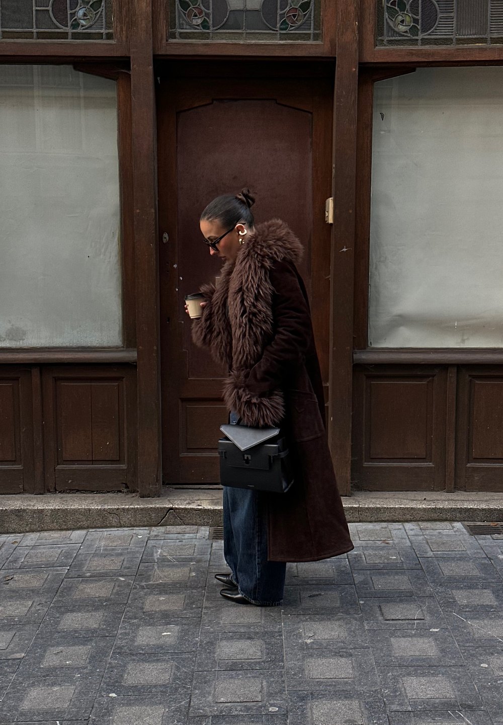 Casaco longo castanho com acabamentos em pele sintética, jeans escuros e uma mala preta, segurando uma chávena de café enquanto caminha numa rua de calçada.