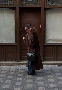 Casaco longo castanho com acabamentos em pele sintética, jeans escuros e uma mala preta, segurando uma chávena de café enquanto caminha numa rua de calçada.