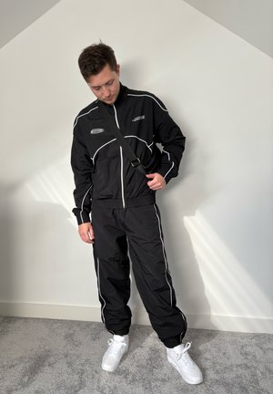 Chándal negro con acentos blancos y chaqueta con cremallera. Presenta puños y cintura elásticos. Combinado con zapatillas blancas sobre una alfombra gris.