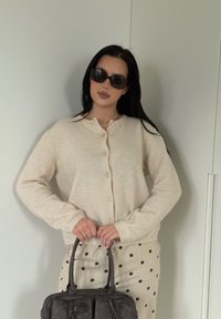 Cardigan beige à col rond et fermeture par boutons, associé à une jupe marron à pois. Porte un sac à main en cuir foncé avec deux poignées.