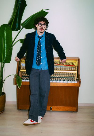 Joven con gafas, chaqueta vaquera, corbata estampada y zapatillas blancas se apoya en un piano vertical abierto junto a una gran planta verde.