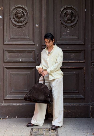 Camisa amarilla claro con botones, pantalones anchos color crema, bolsa de ante marrón oscuro con asa, de pie cerca de una puerta de madera marrón con características ornamentales.