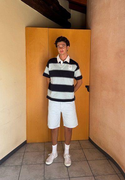 Camisa polo a rayas con cuello blanco, con rayas horizontales en azul marino y azul claro. Combinada con pantalones cortos claros y zapatillas blancas.