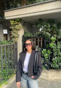 Μαύρο oversized σακάκι πάνω από ένα λευκό πουκάμισο, με ανοιχτό μπλε τζιν. Μια μαύρη τσάντα είναι κρεμασμένη σε έναν ώμο. Πράσινα φυτά και ένας γκρίζος τοίχος είναι στο φόντο.