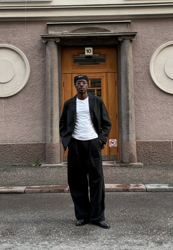 Homme portant un blazer noir, une chemise blanche, un pantalon large noir, une casquette noire et des lunettes, debout dans la rue devant une porte marron avec le numéro 10.
