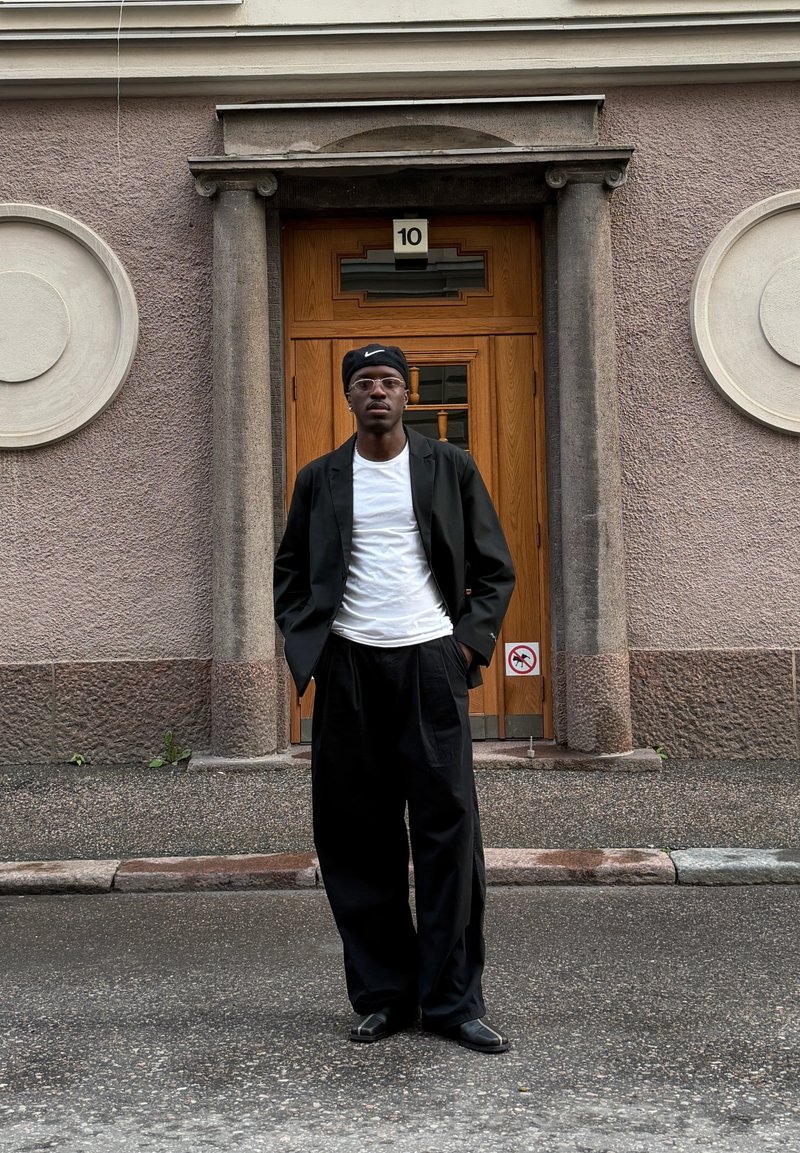 Homme portant un blazer noir, une chemise blanche, un pantalon large noir, une casquette noire et des lunettes, debout dans la rue devant une porte marron avec le numéro 10.