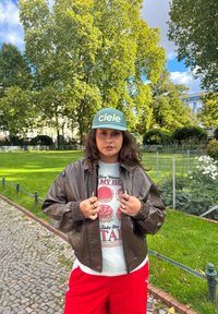 Brun bomberjakke i kunstlæder, grafisk t-shirt i lys grå, røde shorts og en grøn kasket med "ciele"-logo. Flere ringe på hænderne.