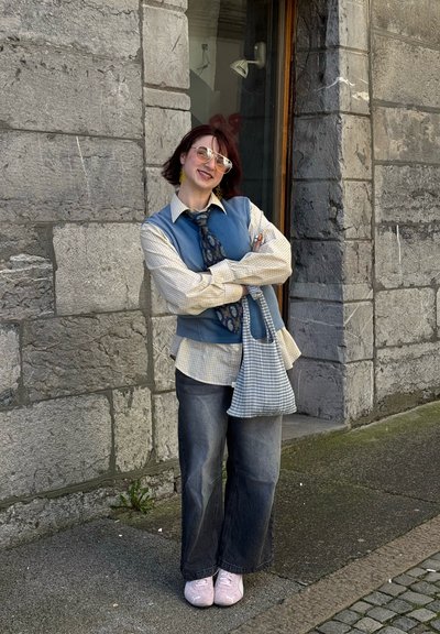 Chaleco azul sobre una camisa de cuadros, atado con una corbata de estampado, combinado con vaqueros oscuros de pierna ancha y zapatillas rosas. Lleva un bolso de cuadros.