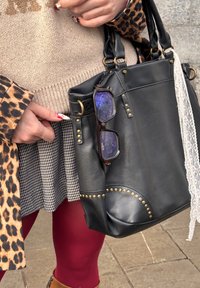 Χέρια που κρατούν μια μαύρη δερμάτινη τσάντα με τρουκς, με γυαλιά, κορδέλα δαντέλας, παλτό με λεοπάρ σχέδιο, καρό φούστα, μπορντό καλσόν και καφέ μπότες.