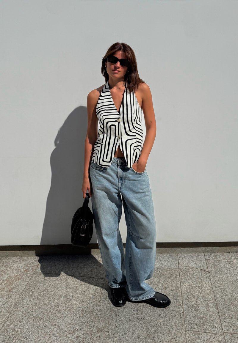 Mulher a usar óculos de sol pretos, top halter geométrico preto e branco, jeans largos azul claro, sapatos pretos, segurando uma mala preta, de pé contra uma parede cinza.