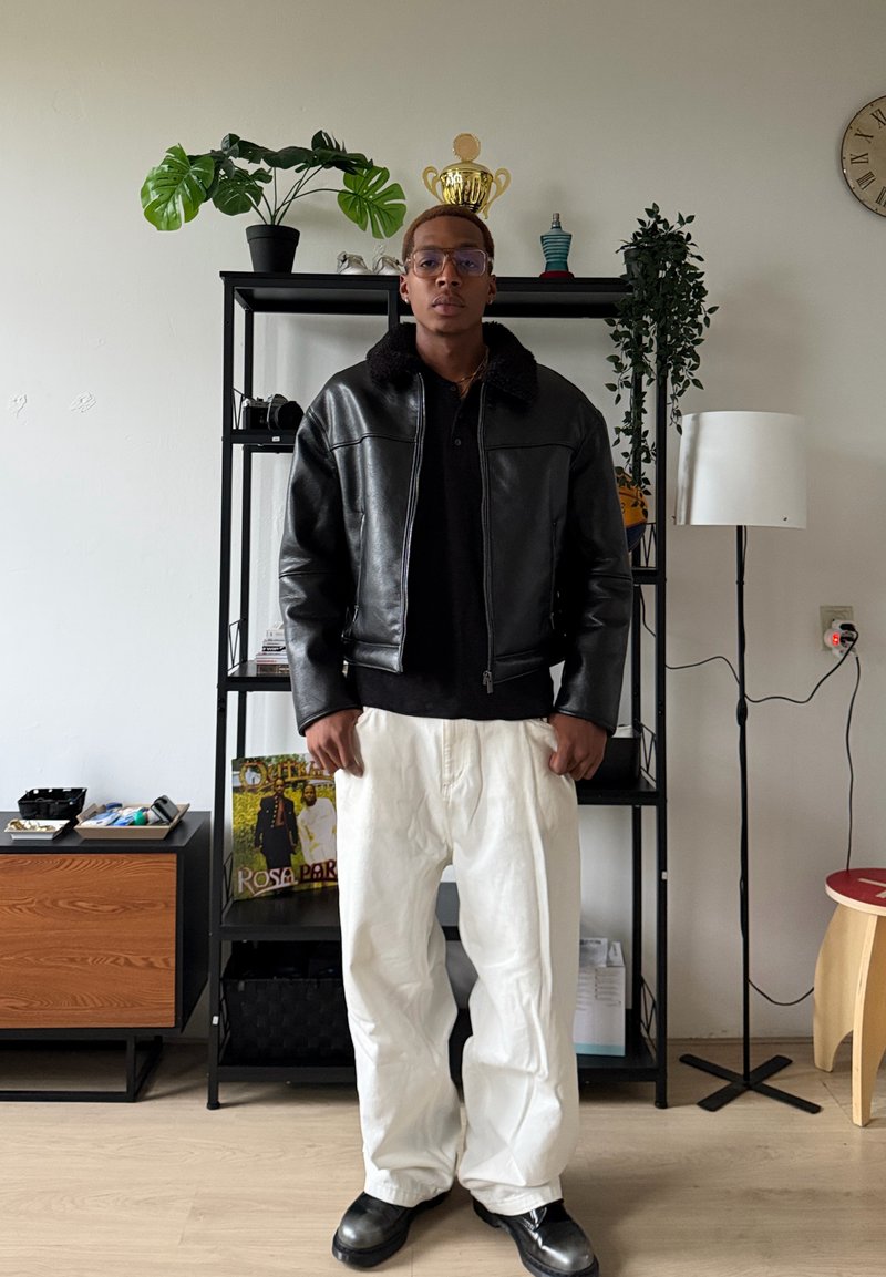 Veste en cuir noire avec col en fausse fourrure, polo noir et pantalon blanc ample ; debout dans une pièce avec des plantes et une étagère.