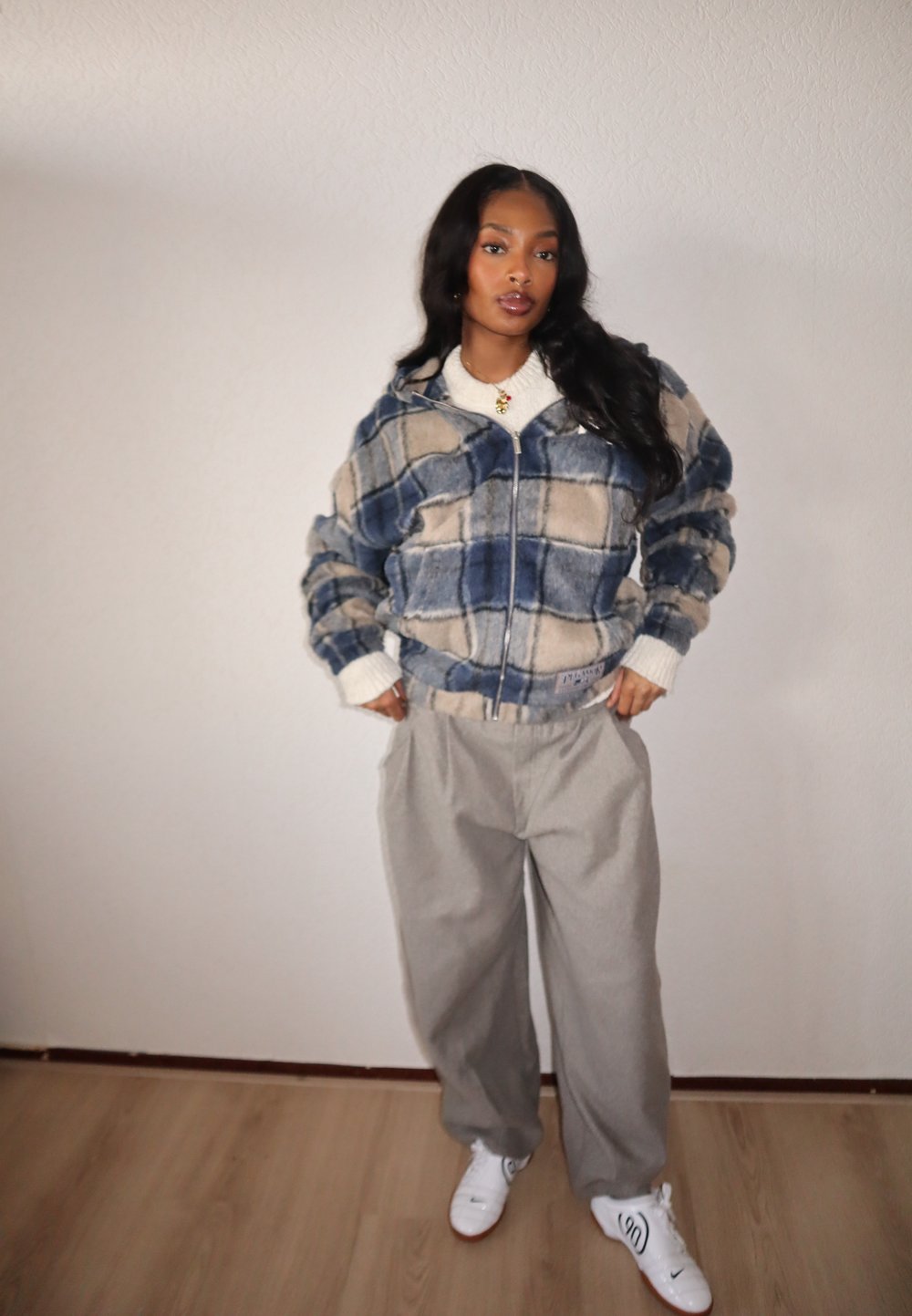 Veste en polaire à carreaux bleu et beige avec une fermeture éclair à l'avant, associée à un pantalon large gris et des baskets blanches. Texture douce, design décontracté.